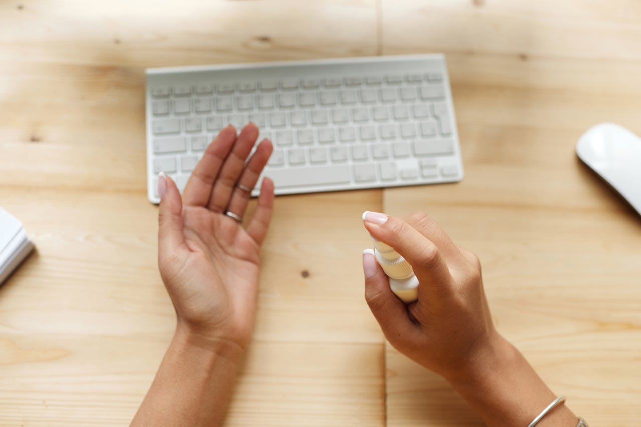 Close-up of hands using sanitizer spray near a keyboard on a wooden desk.